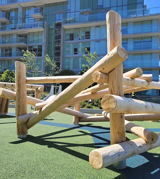 Peeled-log climbing structure on green rubber surfacing in front of a glass apartment building