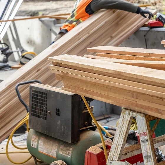 Stacked cedar boards on a sawhorse beside an air compressor at a job site