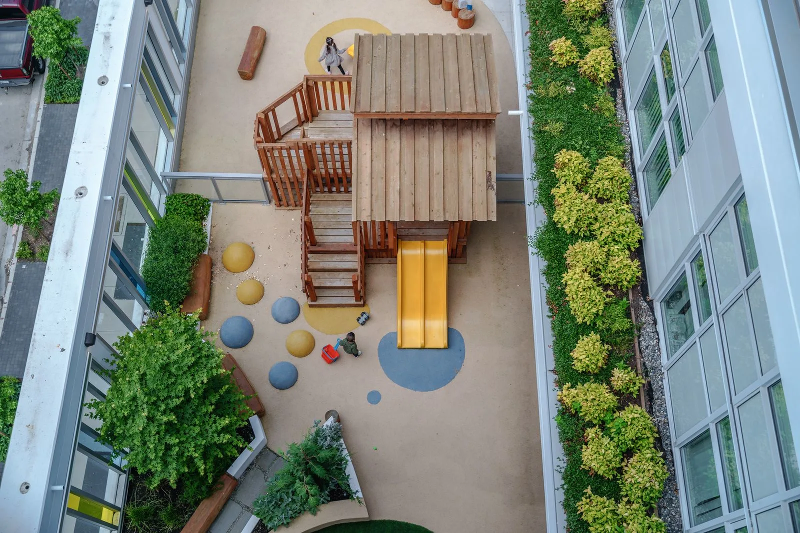 Overhead view of a courtyard playground with a wooden playhouse, yellow slide and coloured ground circles