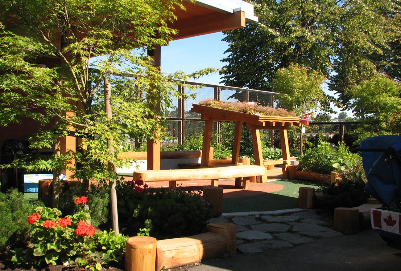 Timber pergola with green roof and log benches in a garden play area beside red flowers