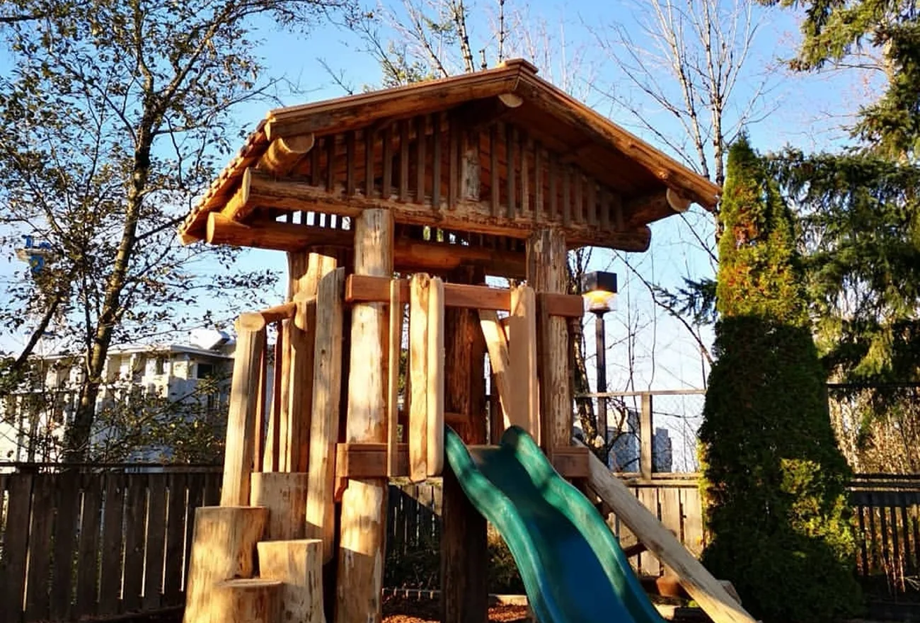 Log playhouse with a shingled roof, wooden steps and a green slide under blue sky