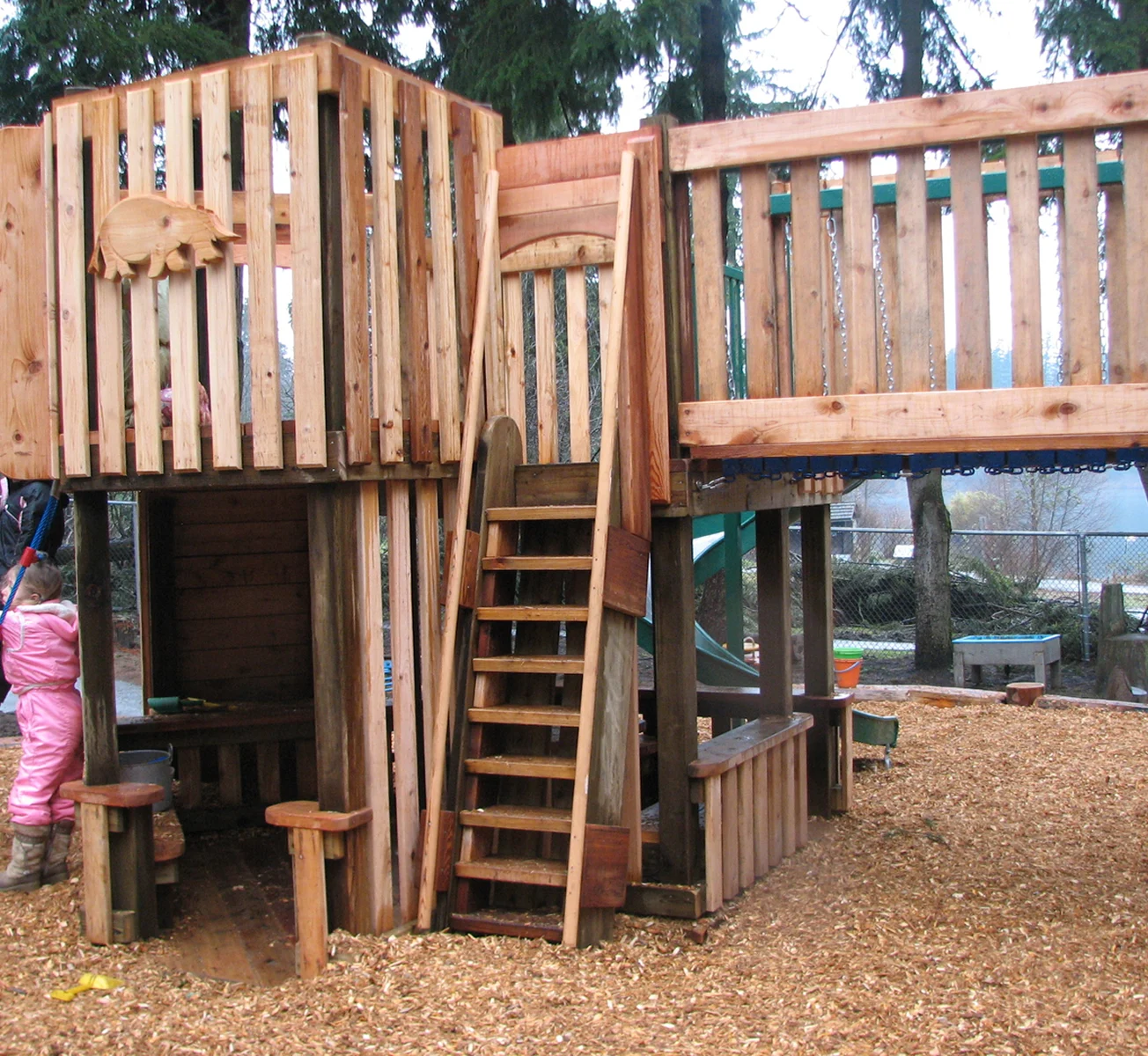 Two-level wooden playhouse with stairs, slatted railings and a carved panel on wood chips
