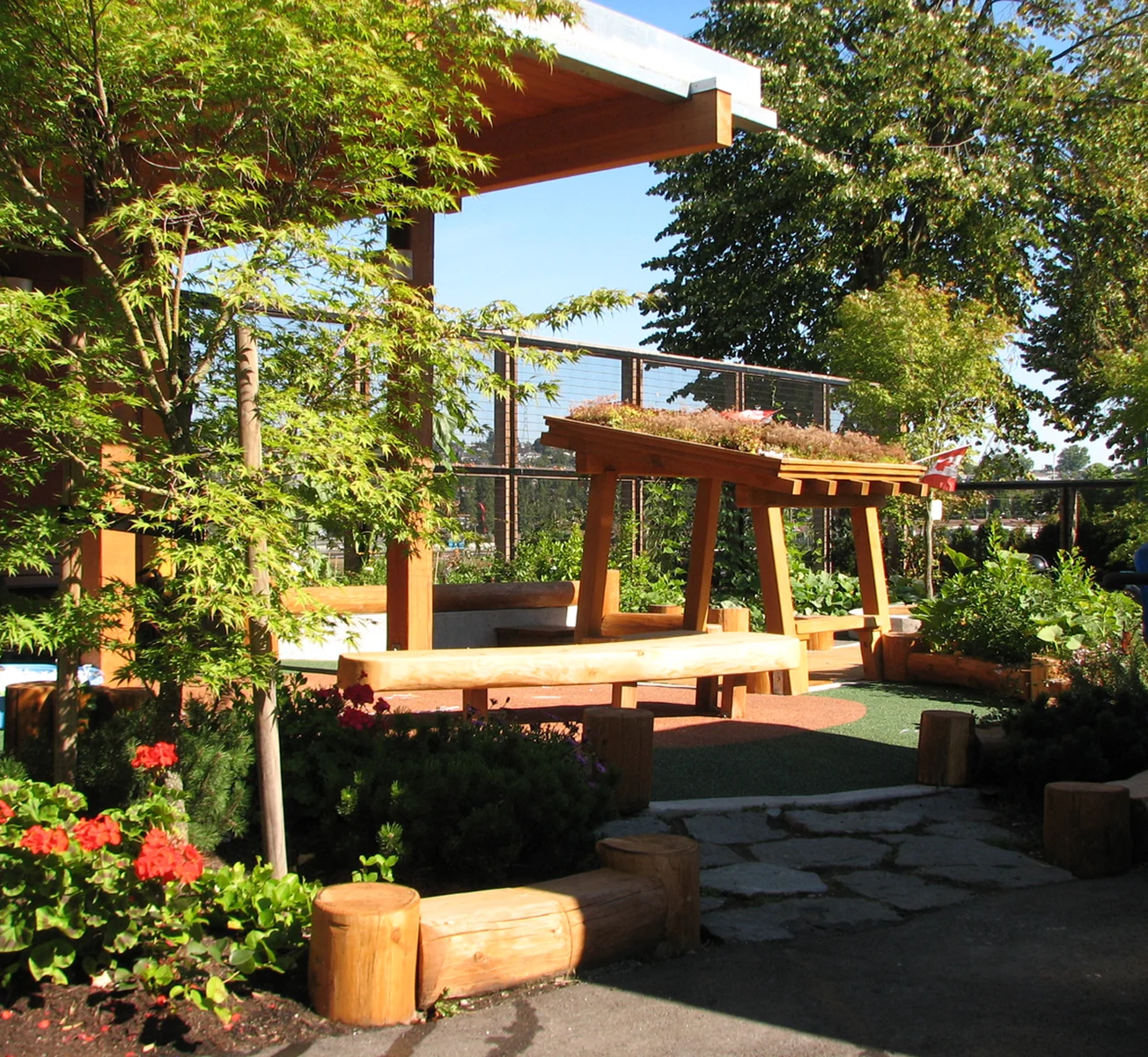Timber pergola with green roof and log benches in a garden play area beside red flowers