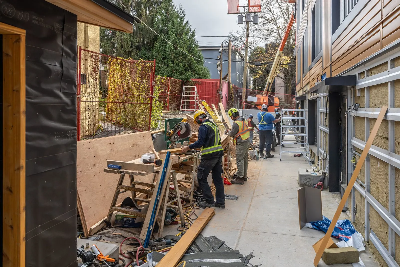 Crew in hard hats and safety vests cutting lumber at a workbench in a construction laneway