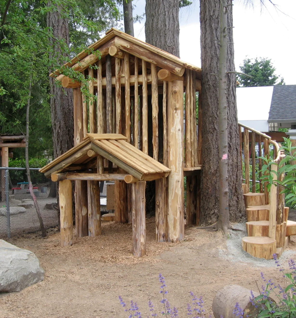 Log cabin playhouse with a covered porch, stump steps and a boulder on sandy ground