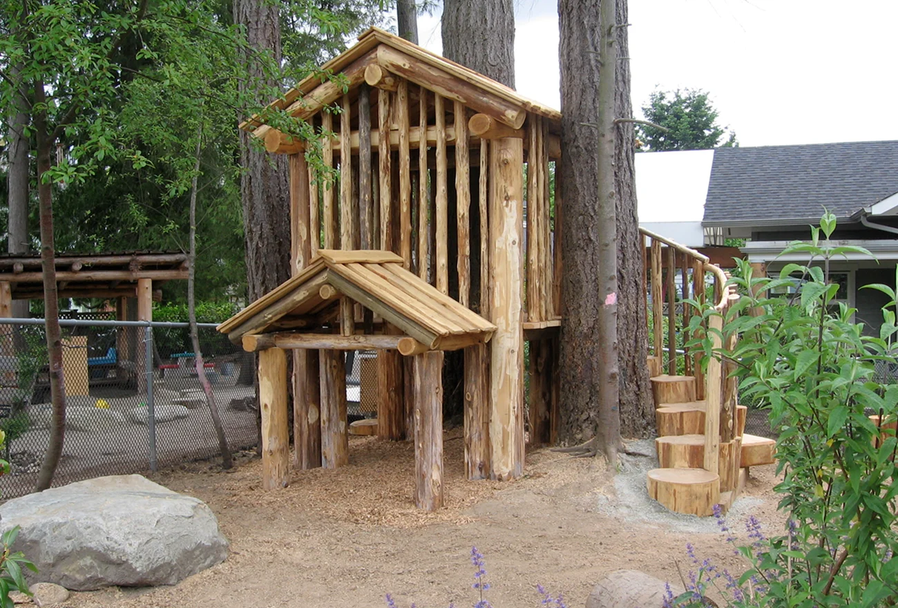 Log cabin playhouse with a covered porch, stump steps and a boulder on sandy ground