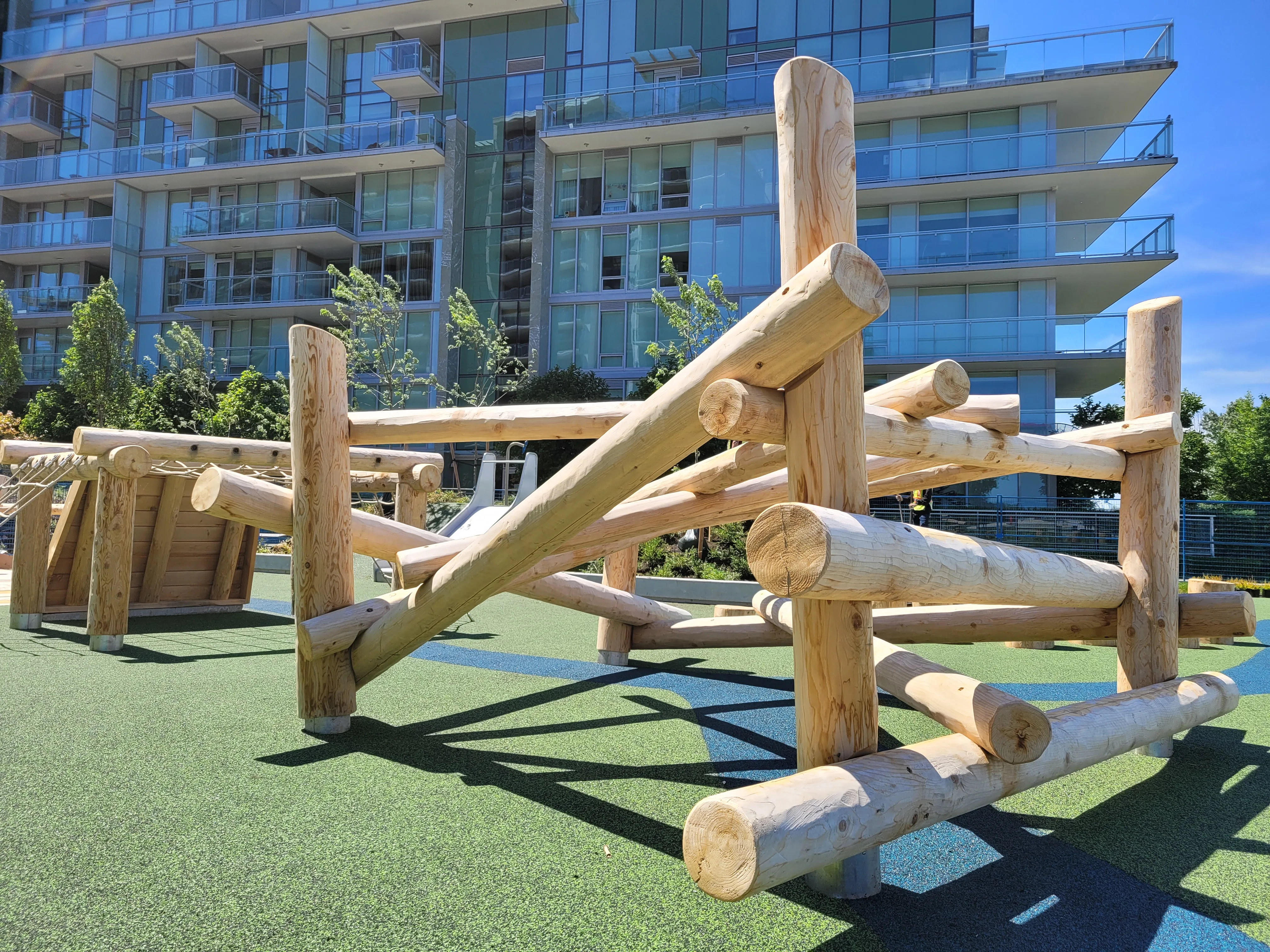 Peeled-log climbing structure on green rubber surfacing in front of a glass apartment building