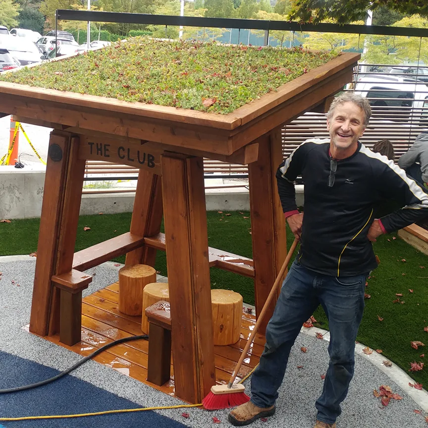 Man standing beside a timber shelter with a planted green roof and stump seats