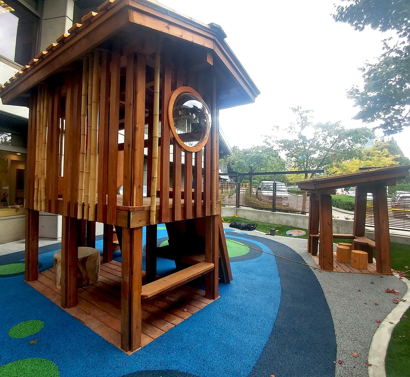 Cedar playhouse with vertical slats and round window on blue rubber surfacing with a pathway