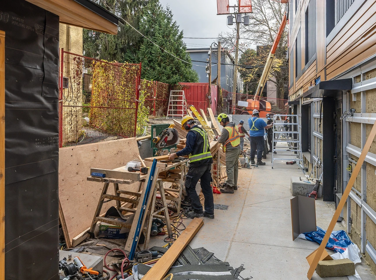Crew in hard hats and safety vests cutting lumber at a workbench in a construction laneway