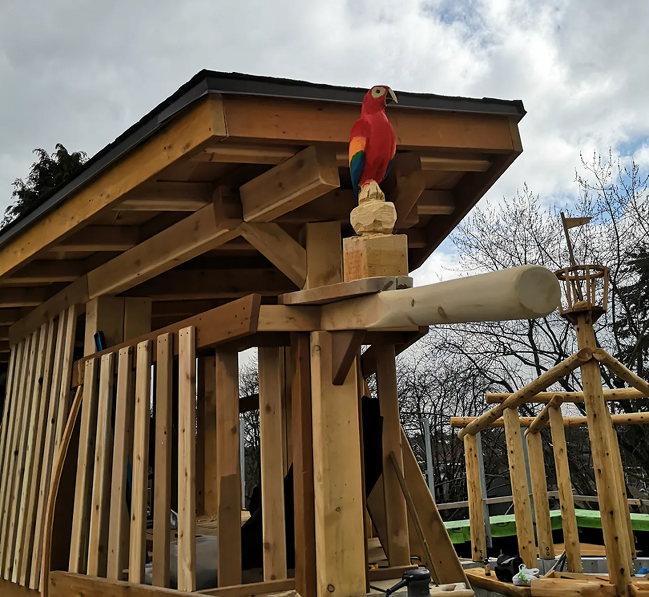 Timber shelter roof topped with a carved red parrot beside a log play structure